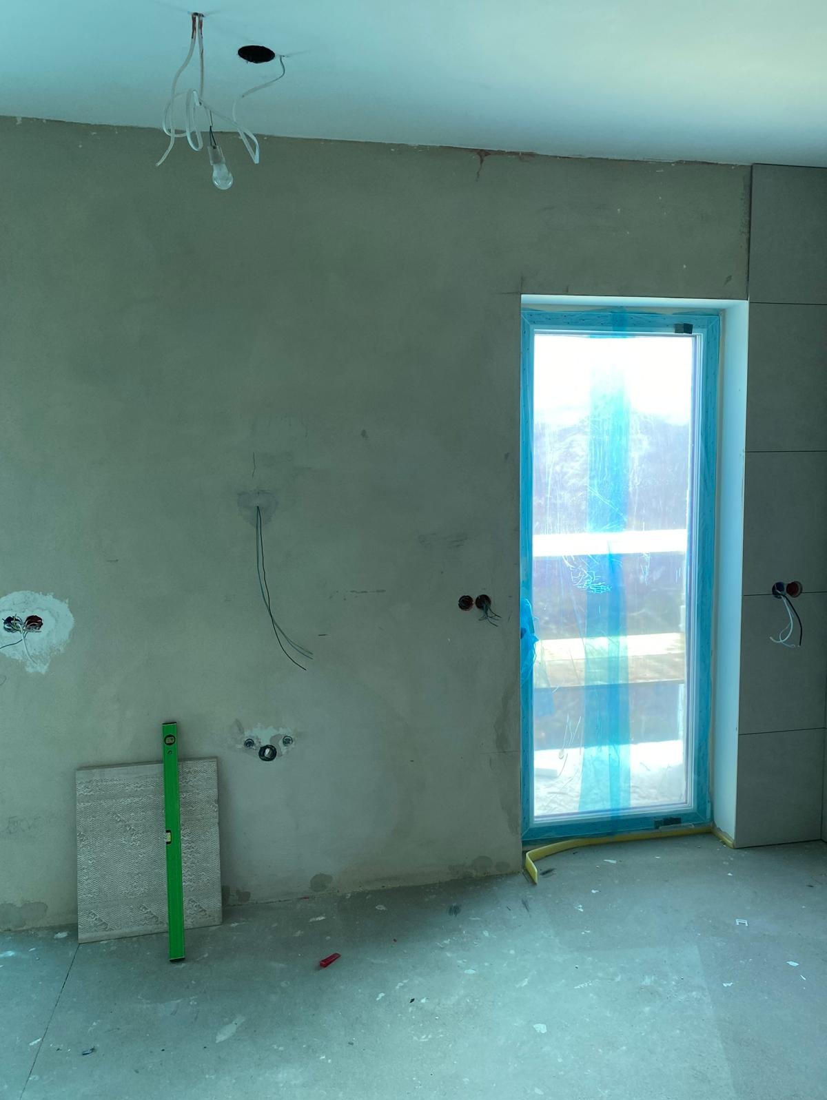 Surowy stan wnętrza w trakcie prac wykończeniowych: ściany po tynkowaniu z widocznymi przewodami elektrycznymi, okno zabezpieczone folią, poziomica oparta o płytę ceramiczną, niezagruntowana podłoga.