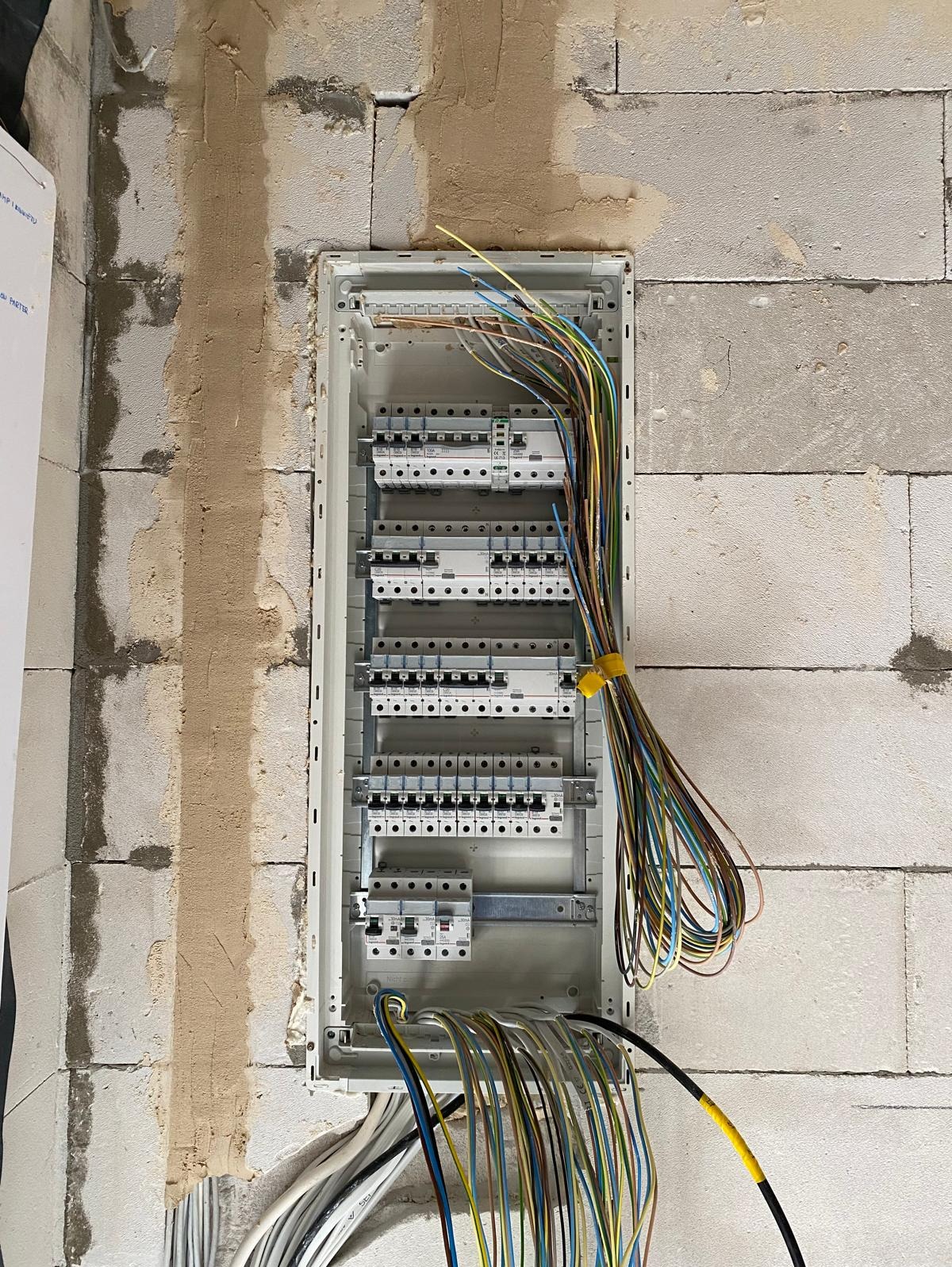 Rozdzielnica elektryczna z podłączonymi przewodami w surowym stanie, widoczne zabezpieczenia i przewody w różnych kolorach, ściana z betonu komórkowego z widocznymi śladami po zaprawie.