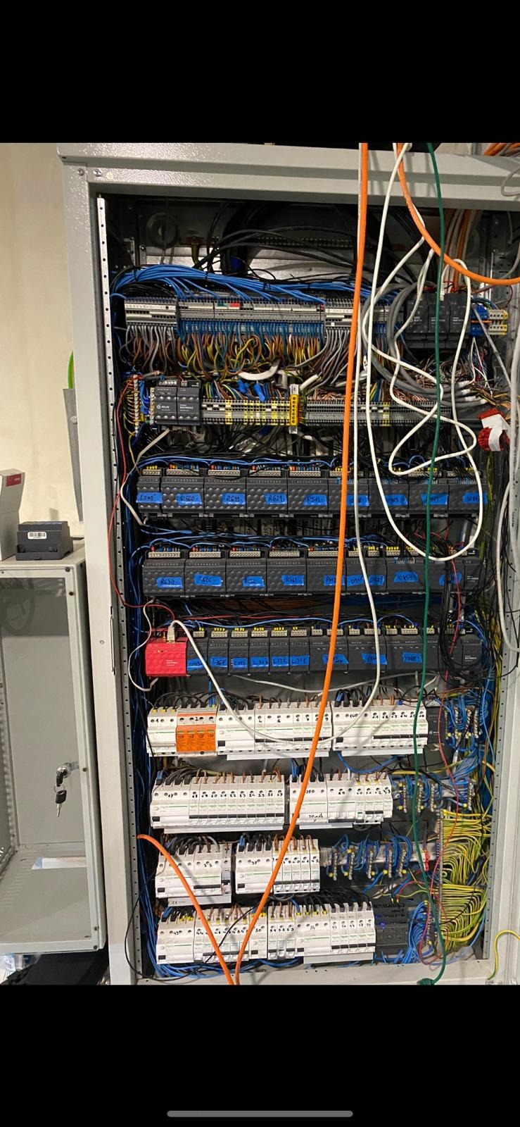 Otwarta szafa sterownicza z widocznymi elementami elektrycznymi, w tym licznymi przewodami w różnych kolorach, modułami sterującymi i zabezpieczeniami; obok szafy widoczny kluczyk i fragment...