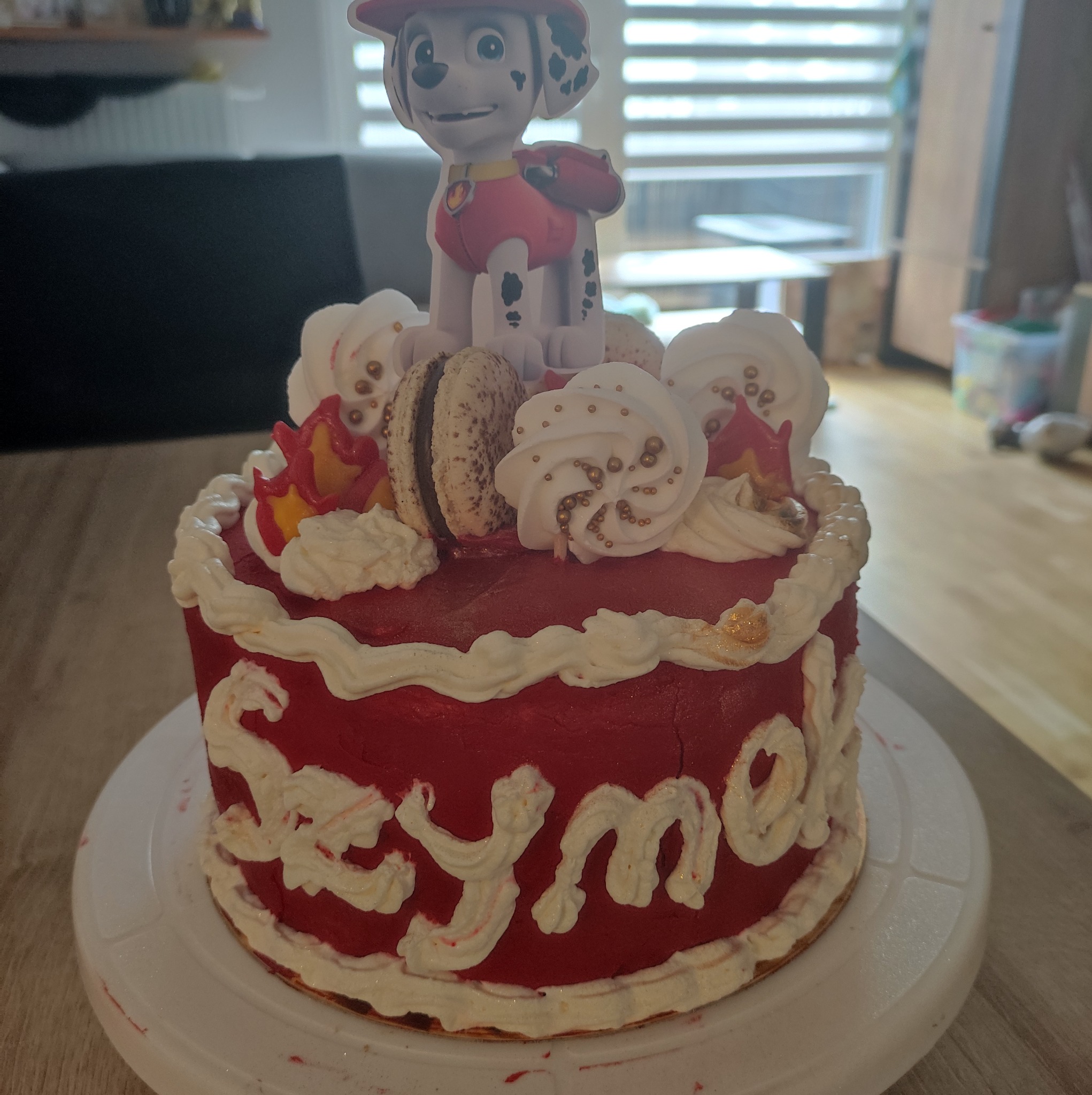 Czerwony tort urodzinowy z białym kremem, makaronikami, dekoracją z motywem bajki Psi Patrol i napisem 'Szymon' wykonanym z kremu.