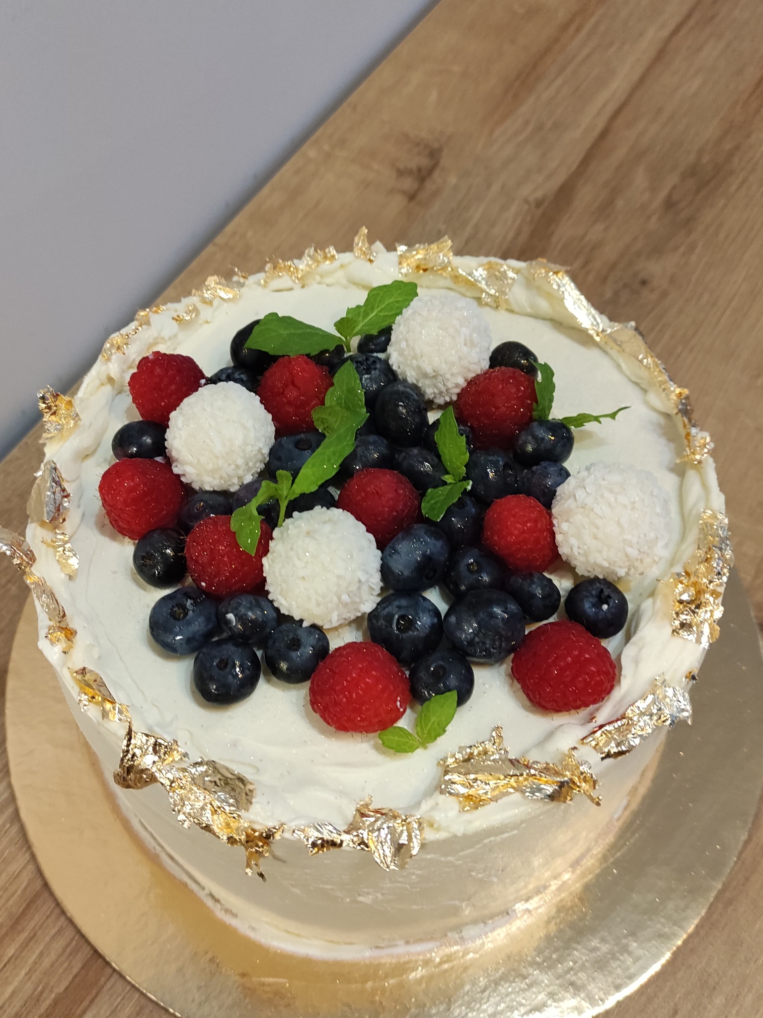 Tort z białym kremem, dekorowany świeżymi borówkami, malinami, kulkami kokosowymi i listkami mięty, z płatkami złota na obrzeżach, na drewnianym blacie.