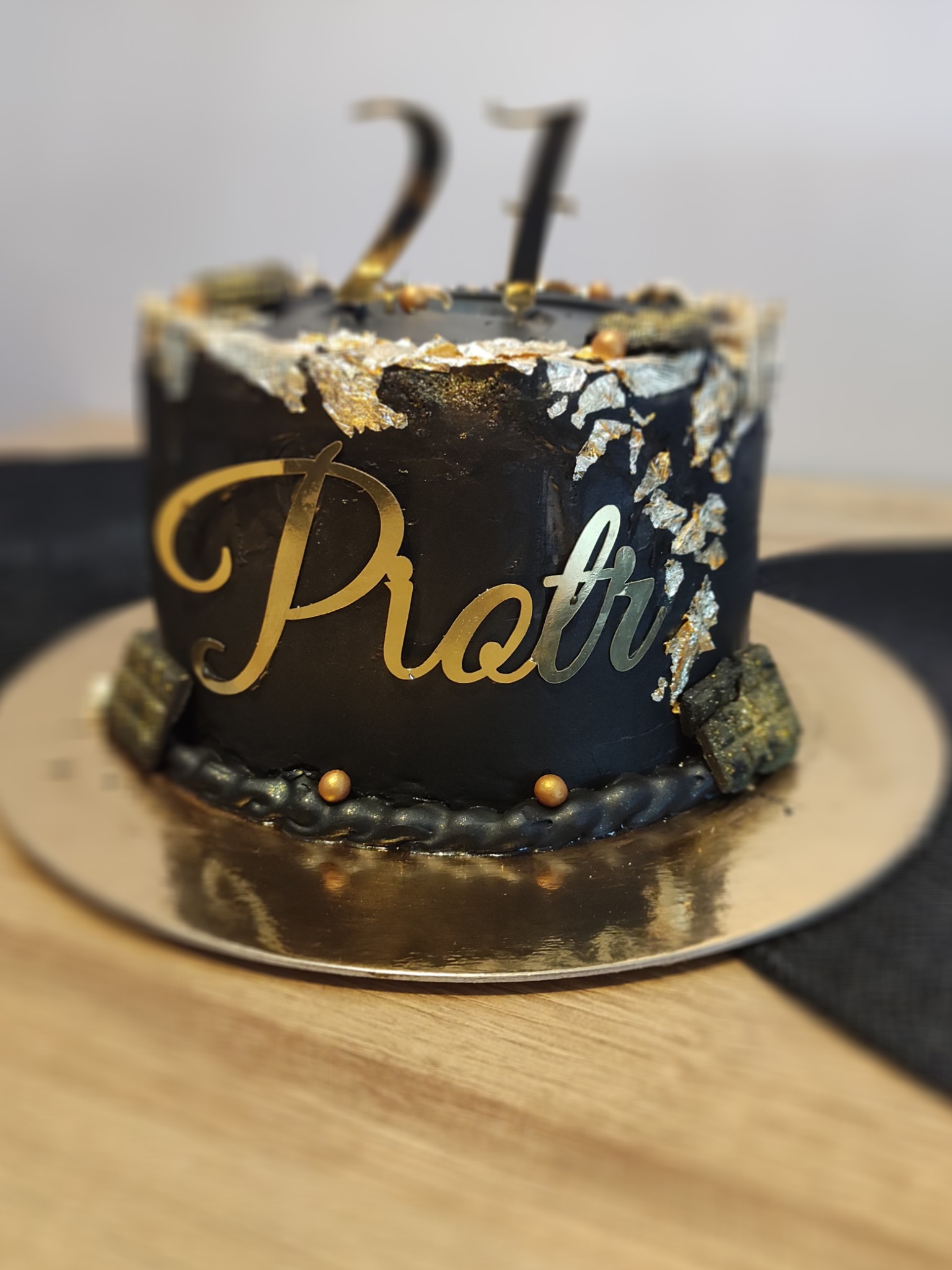 Czarny tort urodzinowy z dekoracją ze złotych płatków, napisem 'Piotr' i cyfrą 27 na złotym podkładzie.
