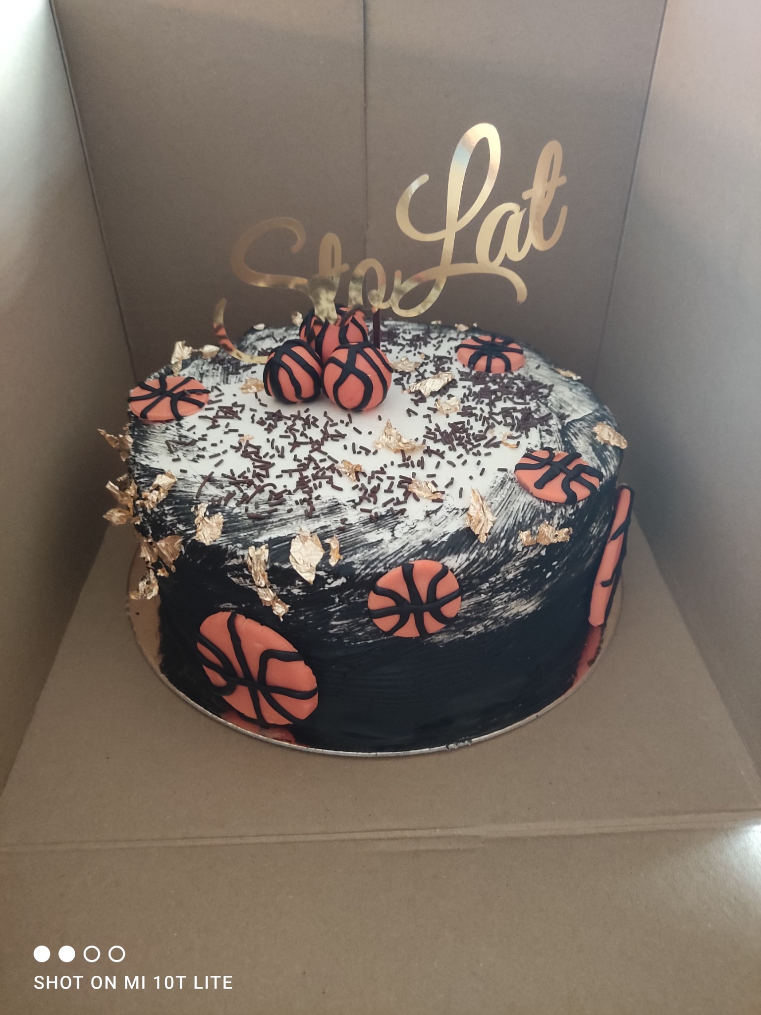 Tort urodzinowy w kartonowym pudełku, dekorowany motywem koszykówki z napisem 'Sto Lat' w złotym kolorze.