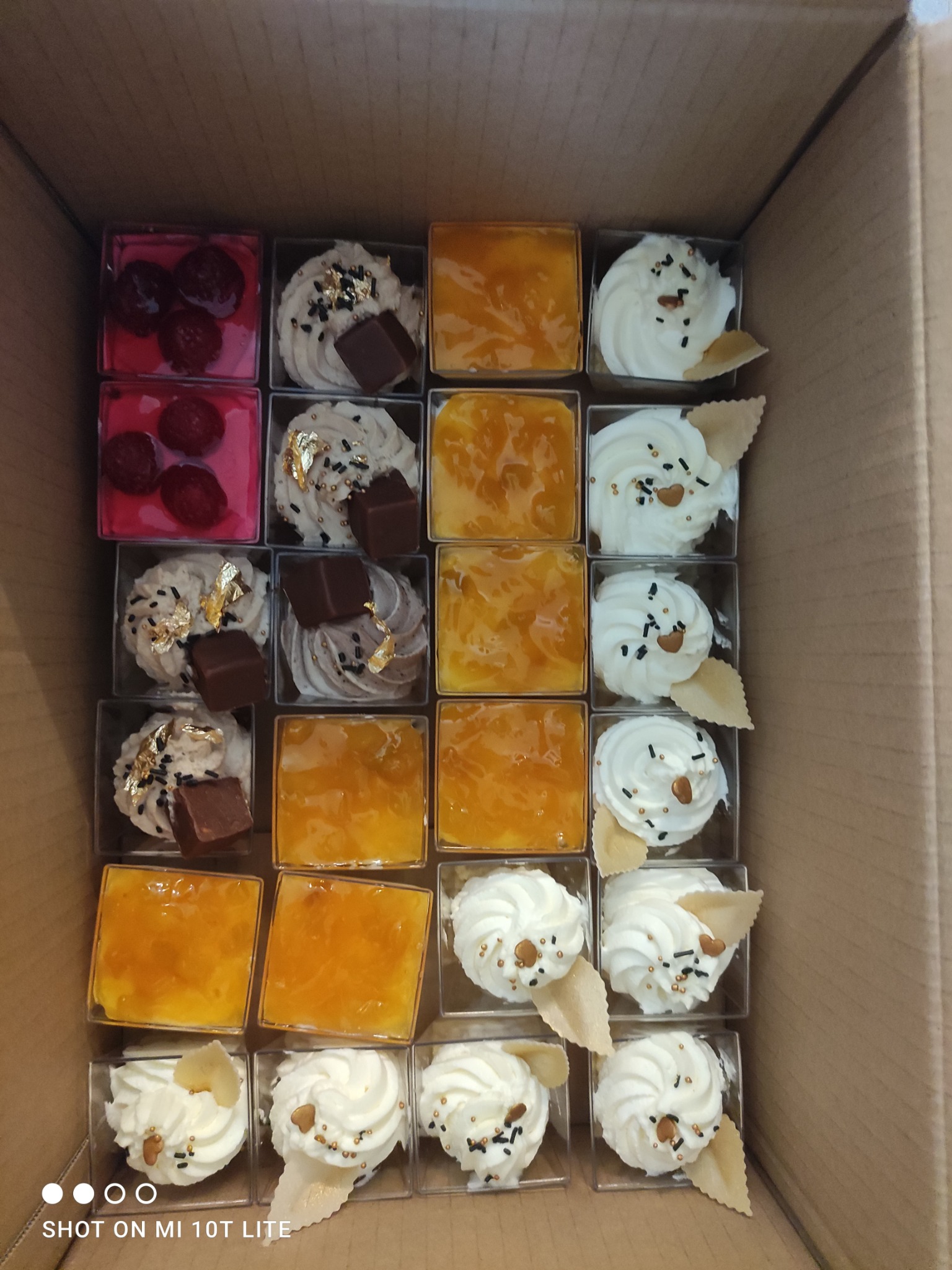 Karton z różnorodnymi deserami w małych, przezroczystych pojemnikach: galaretka malinowa, krem z czekoladą i złotymi płatkami, deser mango i krem z dekoracją z wafelka.