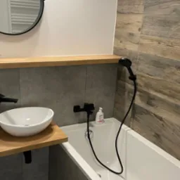 Nowoczesna łazienka z drewnopodobnymi płytkami na ścianie, białą umywalką nablatową z czarną armaturą, białą wanną i okrągłym lustrem z czarną ramą.