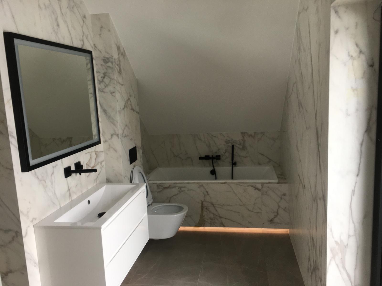 Wykończona łazienka na poddaszu z marmurowymi płytkami na ścianach, białą umywalką z szafką, podświetlaną podłogą i czarną armaturą.