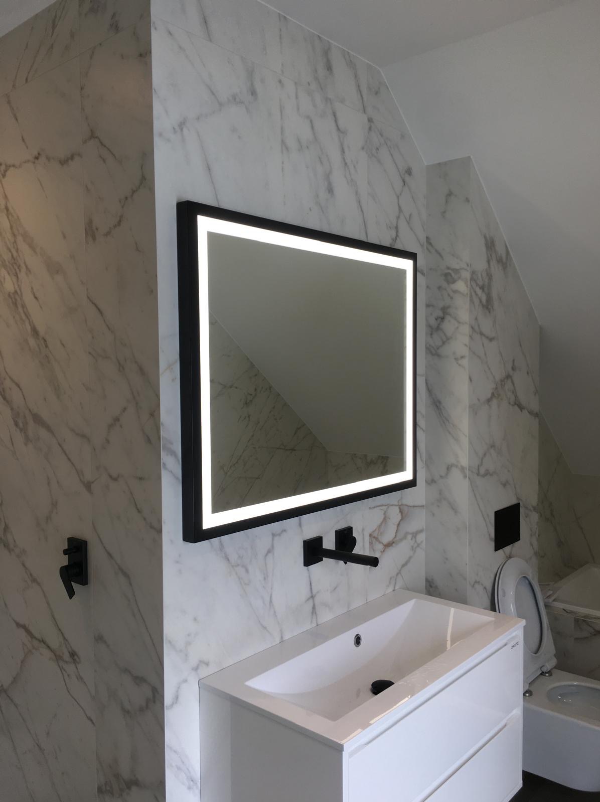 Nowoczesna łazienka z marmurowymi płytkami, podświetlanym lustrem w czarnej ramie, białą umywalką z szafką i czarną armaturą.
