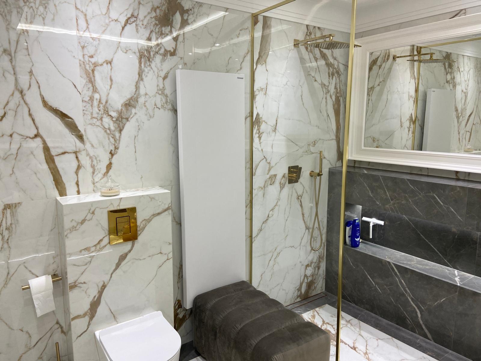 Elegancka łazienka z marmurowymi płytami w odcieniach bieli, brązu i szarości, złote akcenty armatury i kabiny prysznicowej, biała ceramika sanitarna i grzejnik, szara pufa przy grzejniku.