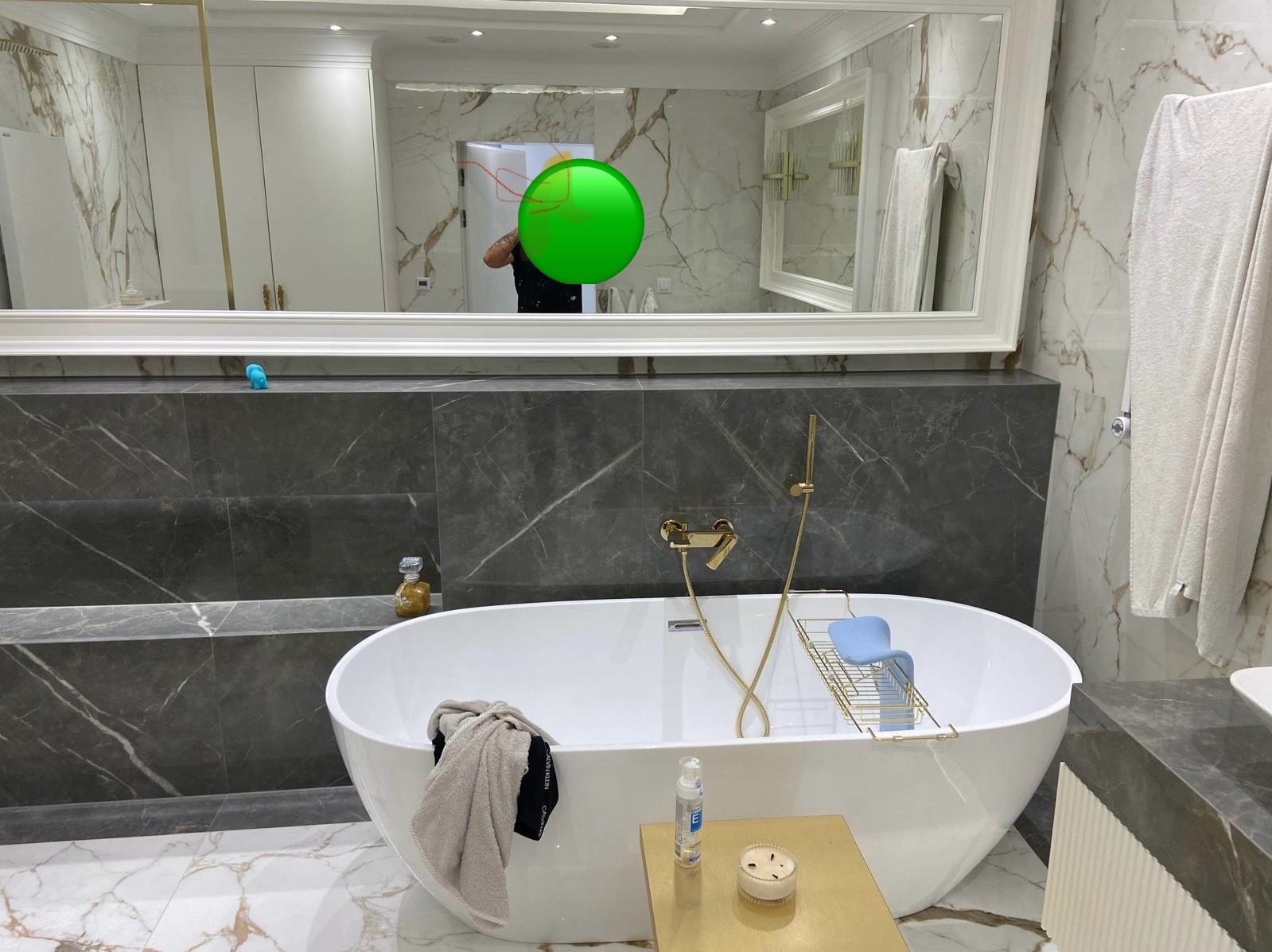 Nowoczesna łazienka z wolnostojącą wanną, marmurowe płytki na ścianach, złota armatura i dodatki, w lustrze odbija się osoba.