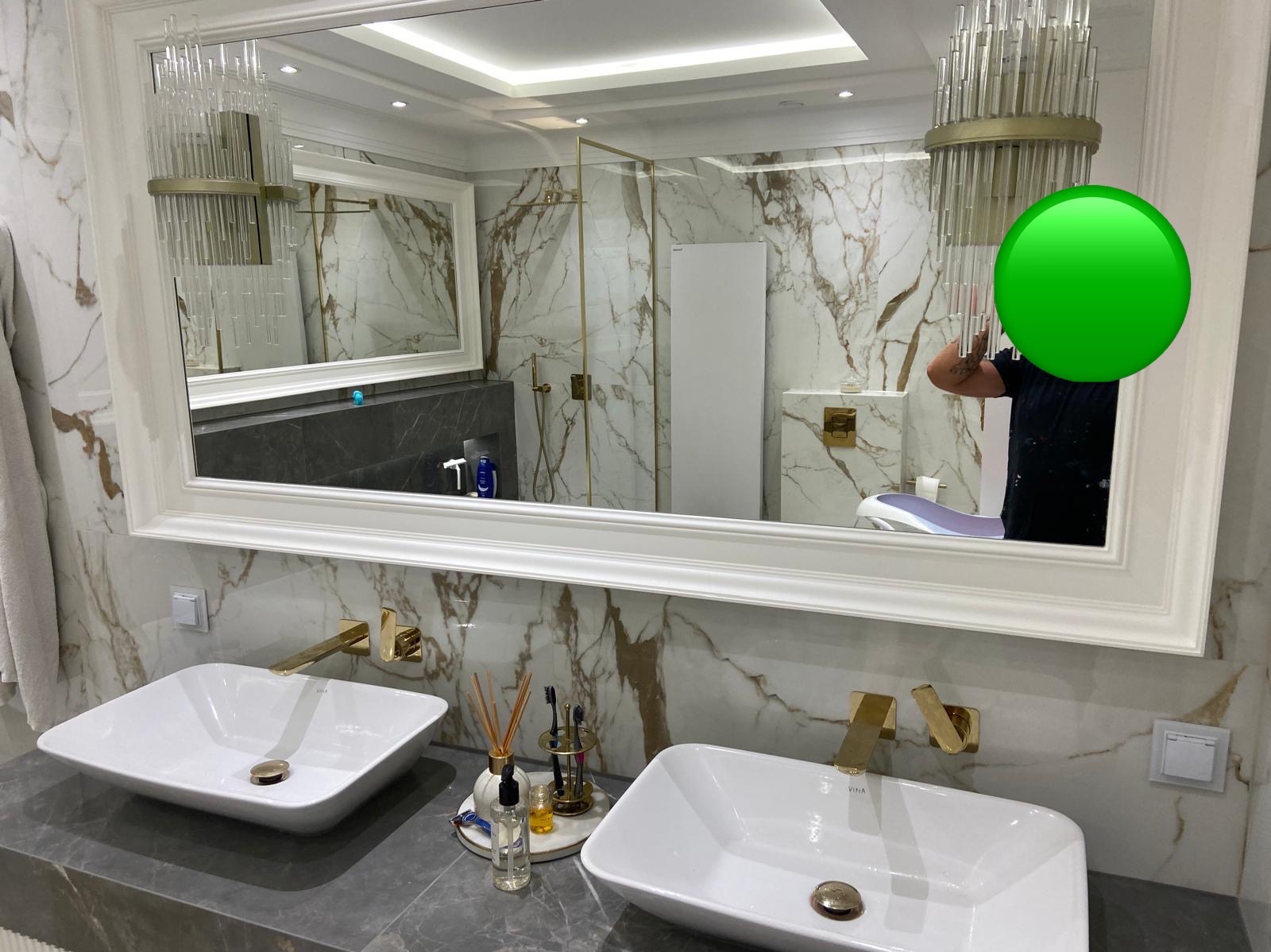 Luksusowa łazienka z dwoma umywalkami nablatowymi, złotą armaturą i marmurowymi płytkami, widoczna w lustrze kabina prysznicowa ze złotymi elementami i ozdobny żyrandol.