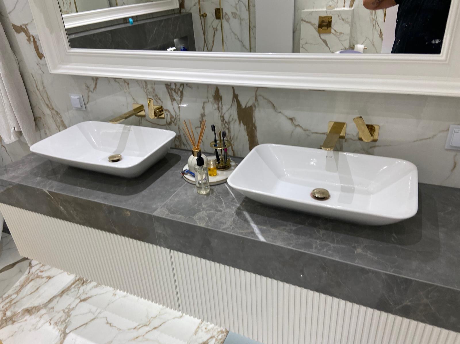 Dwie białe, prostokątne umywalki nablatowe z polerowanego kamienia, złote baterie ścienne, marmurowe ściany i podłoga z widocznym fragmentem szafki z białymi, żłobionymi frontami.