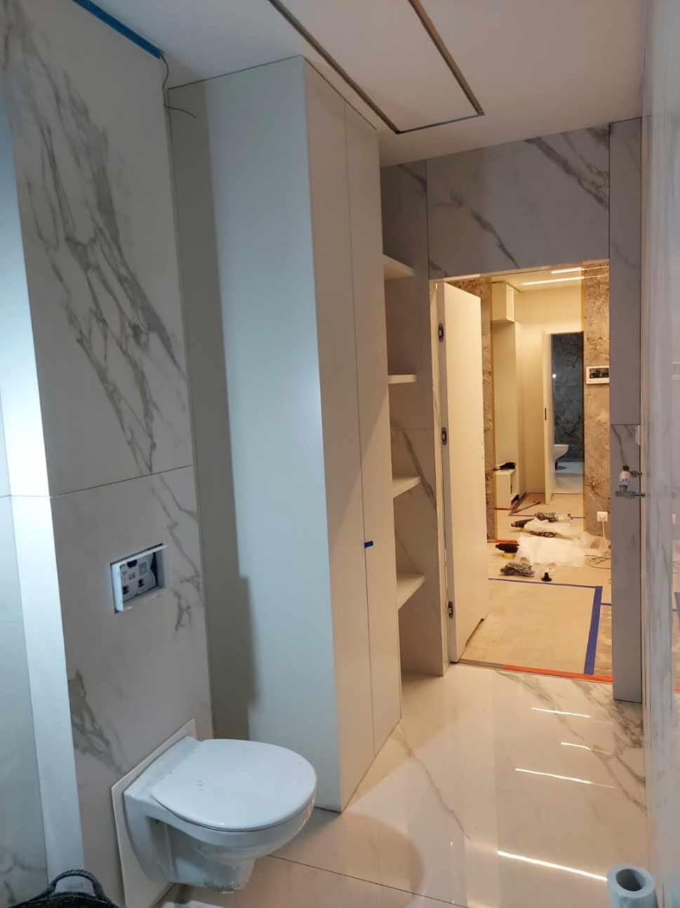 Nowoczesna łazienka w trakcie wykańczania, widoczne marmurowe płytki na ścianach, podwieszana toaleta, biała szafa w zabudowie i otwarte drzwi do kolejnego pomieszczenia z widocznym zabezpieczonym...