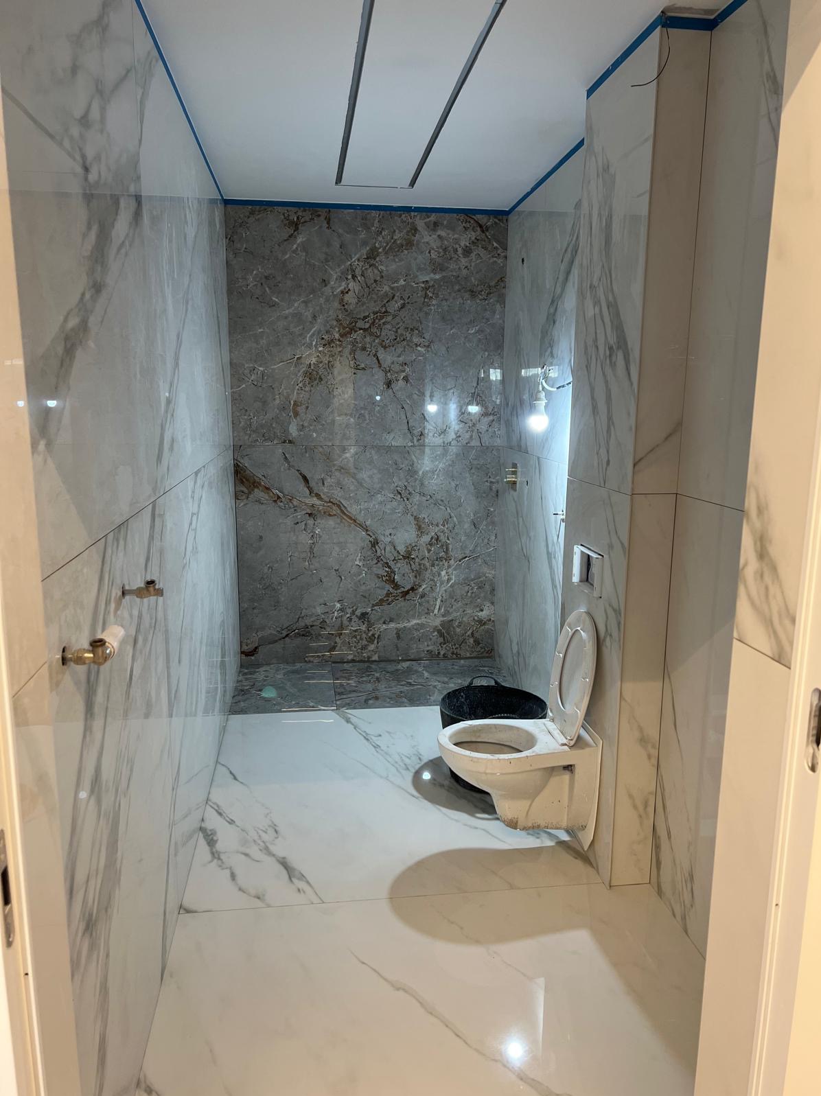 Wnętrze łazienki w trakcie remontu, widoczne płytki imitujące marmur na ścianach i podłodze, niezabudowana instalacja wodna, wisząca toaleta przed montażem, niebieska taśma malarska na obrzeżach...