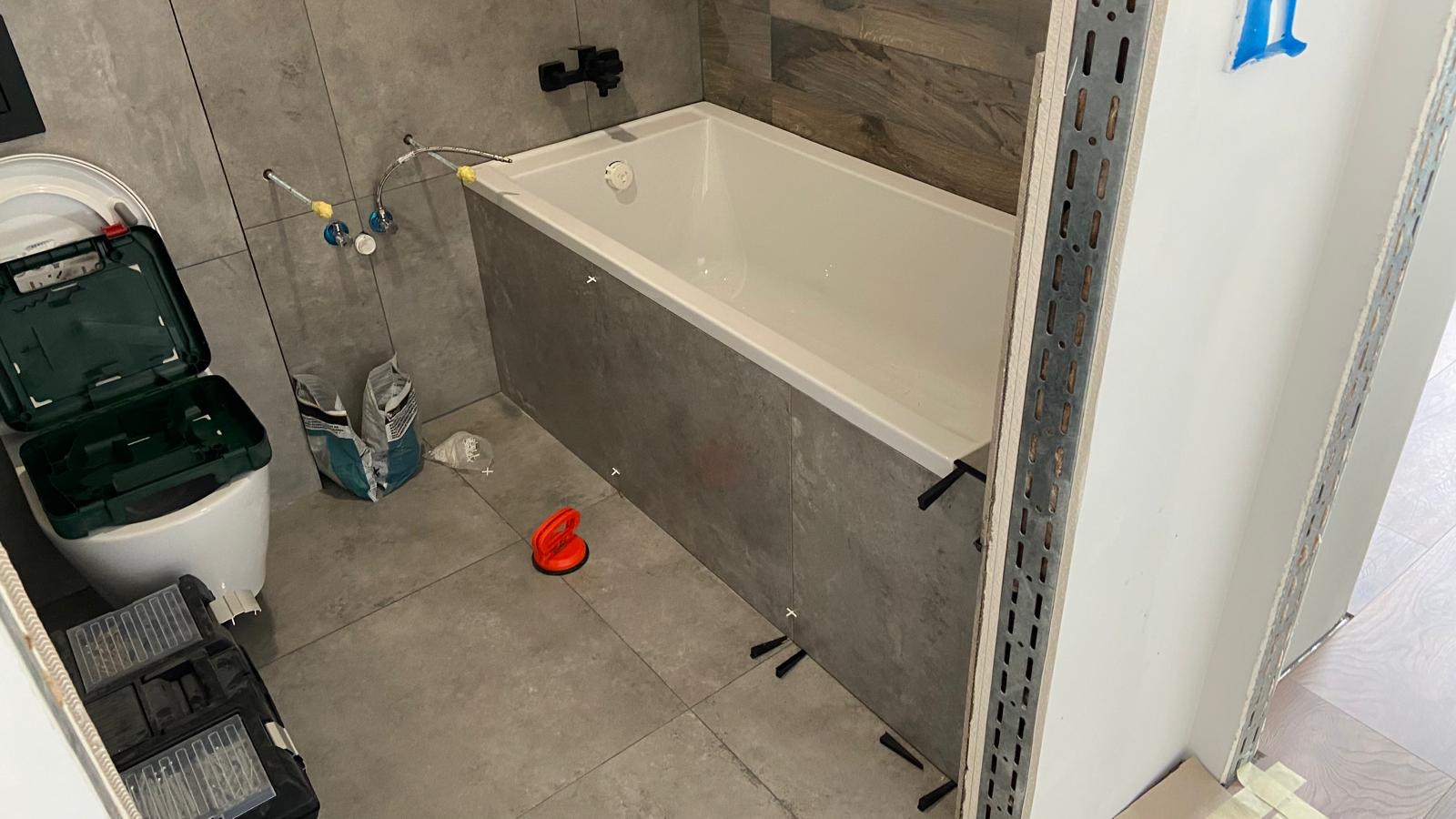 Wnętrze łazienki w trakcie prac: obudowana wanna z szarą okładziną, narzędzia glazurnicze, worki z zaprawą i widoczne elementy instalacji wodnej.