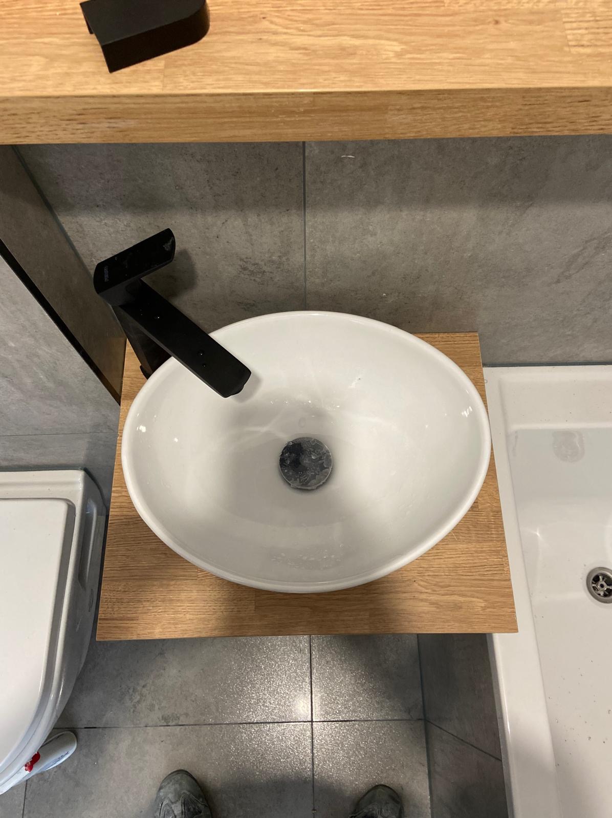 Owalna, biała umywalka nablatowa z czarną, minimalistyczną baterią, zamontowana na drewnianym blacie w łazience wykończonej szarymi płytkami, widok z góry.