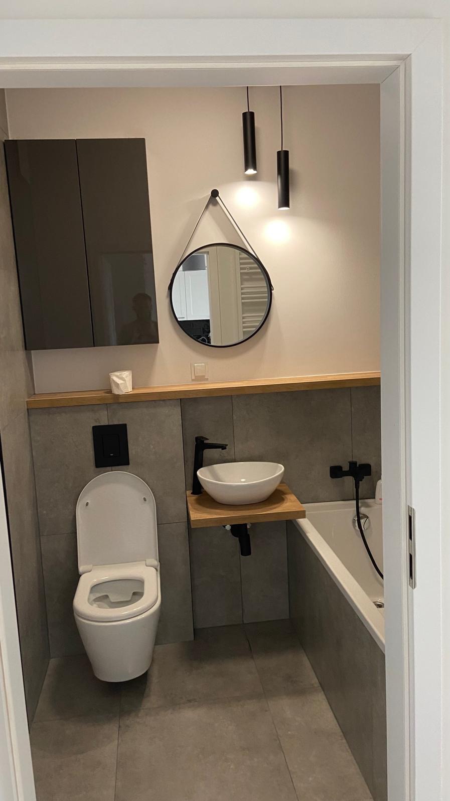 Nowoczesna łazienka w szarościach z drewnianymi akcentami: wisząca toaleta, okrągłe lustro na sznurku, umywalka nablatowa na drewnianym blacie, czarne lampy tuby.