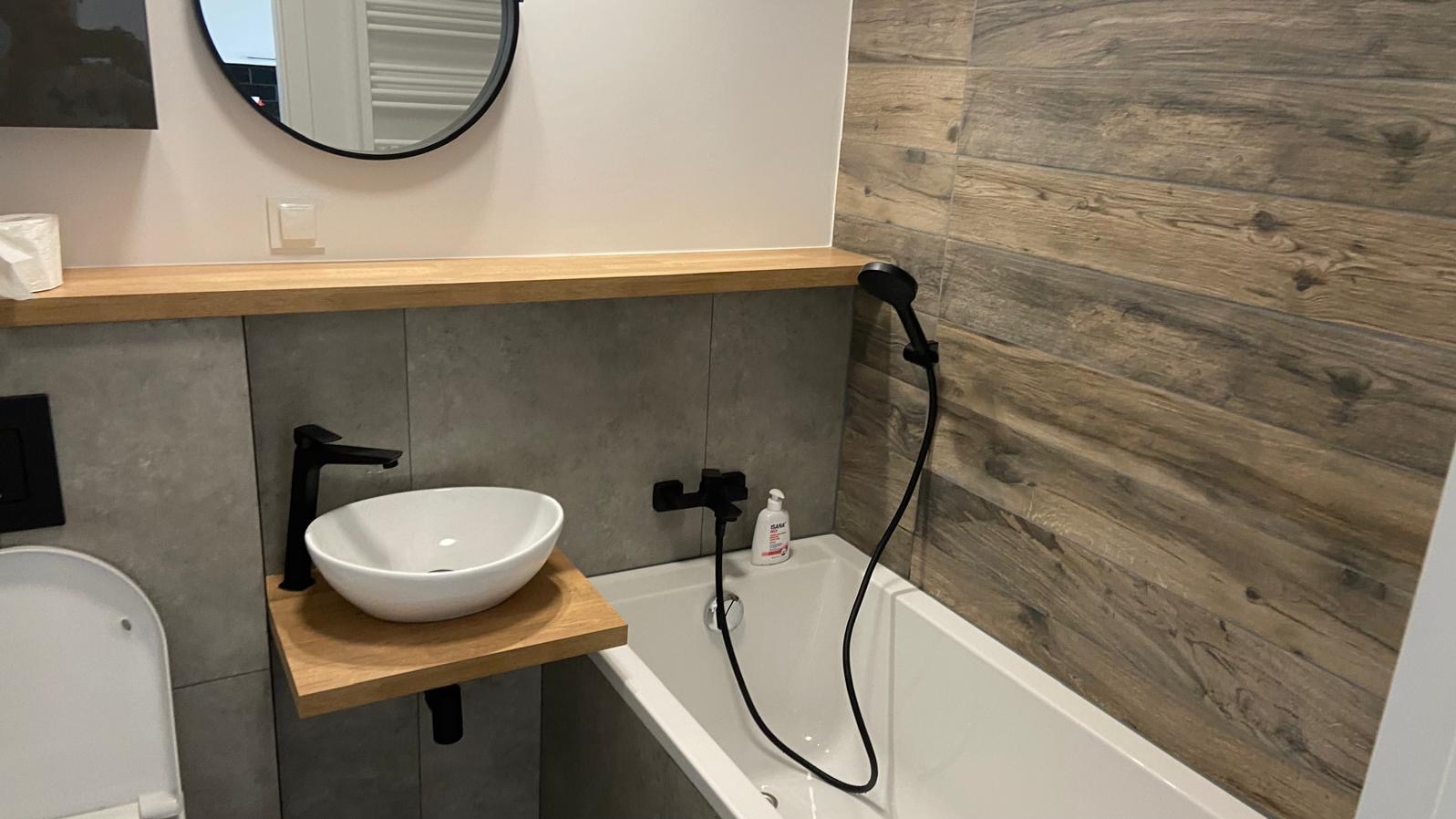 Nowoczesna łazienka z drewnopodobnymi płytkami na ścianie, białą umywalką nablatową z czarną armaturą, białą wanną i okrągłym lustrem z czarną ramą.