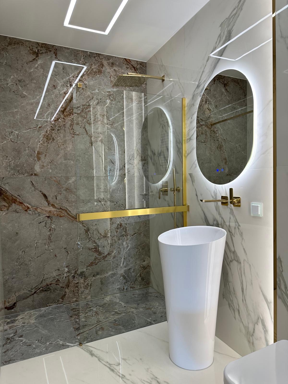 Nowoczesna łazienka z marmurowymi płytkami, kabiną prysznicową typu walk-in, białą umywalką wolnostojącą i podświetlanym lustrem owalnym.
