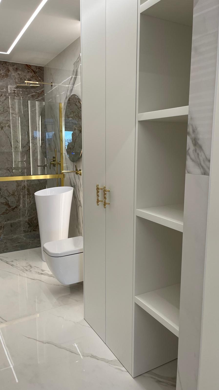 Nowoczesna łazienka z marmurowymi ścianami, wolnostojącą umywalką, wiszącą toaletą i zabudową meblową z ozdobnymi, złotymi uchwytami.