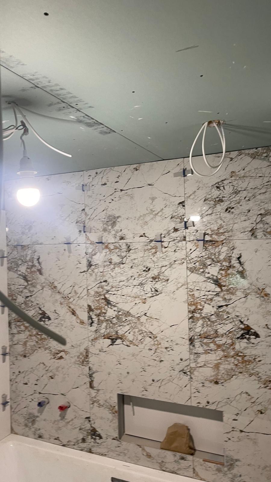 Montaż płytek imitujących marmur w łazience, widoczne krzyżyki dystansowe, surowy sufit z instalacją elektryczną, przygotowane podejścia wodne i wnęka w ścianie.