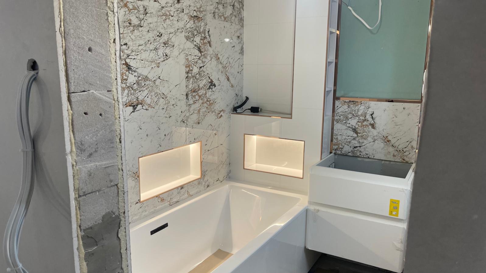 Niedokończona łazienka z wanną, marmurowymi płytkami i podświetlanymi niszami, widoczne surowe ściany i instalacja elektryczna.