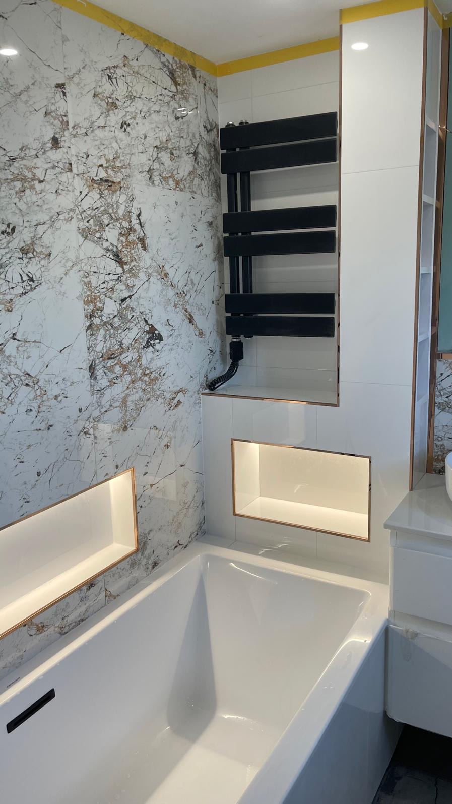 Nowoczesna łazienka z wanną, marmurowymi płytkami i czarnym grzejnikiem drabinkowym, podświetlane wnęki w ścianie z miedzianymi ramkami.
