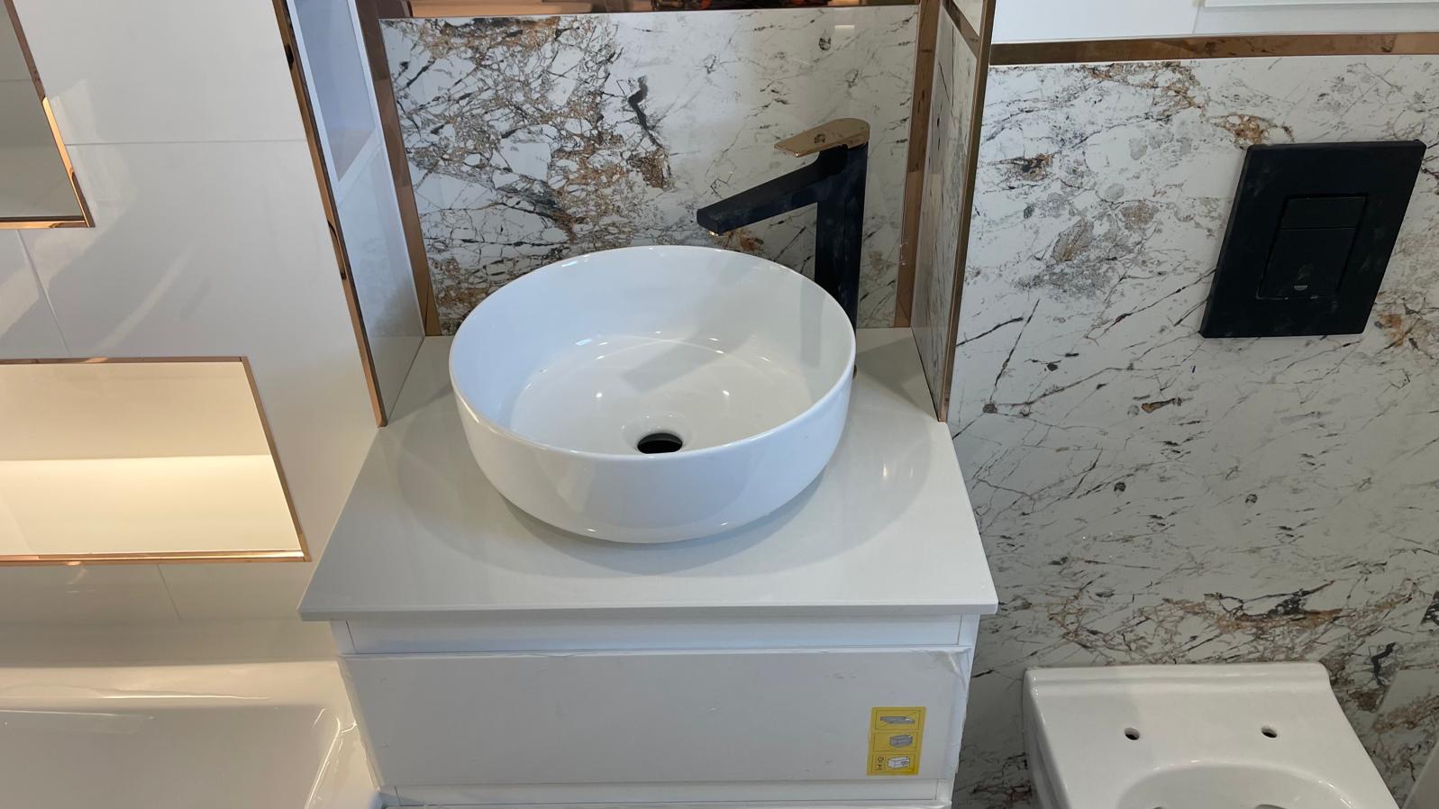 Biała, okrągła umywalka nablatowa z czarną baterią na tle marmurowej ściany z czarnym przyciskiem spłuczki, całość w nowoczesnej łazience z elementami w kolorze miedzianym.