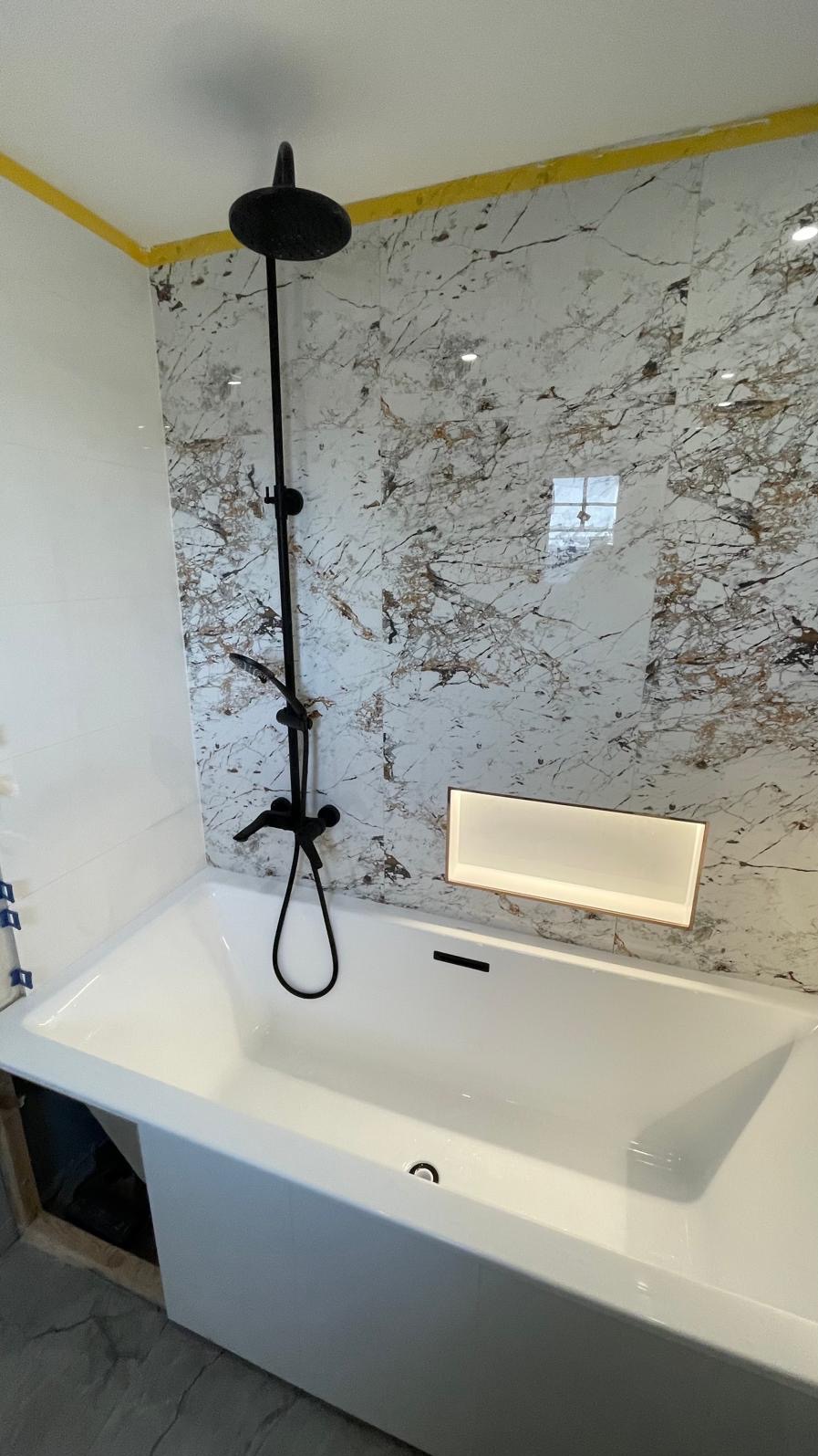 Biała wanna z czarną armaturą i deszczownicą na tle ściany wyłożonej marmurkowymi płytkami, podświetlana półka w ścianie, żółte wykończenie sufitu.
