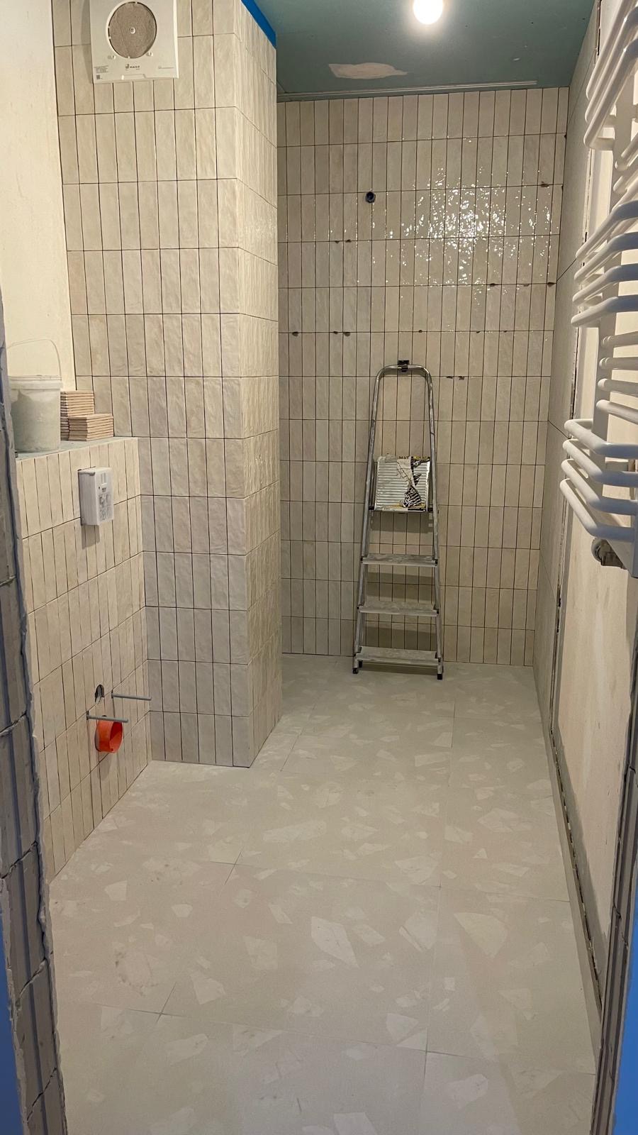 Wnętrze łazienki w trakcie remontu: pionowo ułożone, beżowe płytki na ścianach, szara podłoga z wzorem terrazzo, drabina oparta o ścianę, widoczne instalacje hydrauliczne.