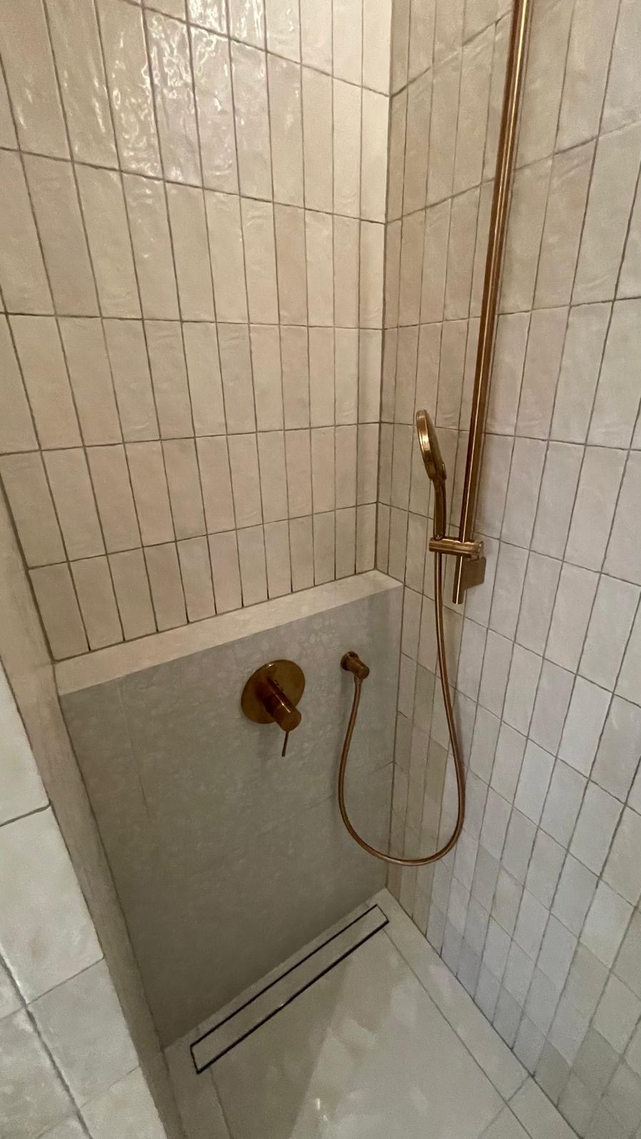 Nowoczesny prysznic z pionowo ułożonymi, kremowymi płytkami, złotą armaturą i odpływem liniowym w posadzce.