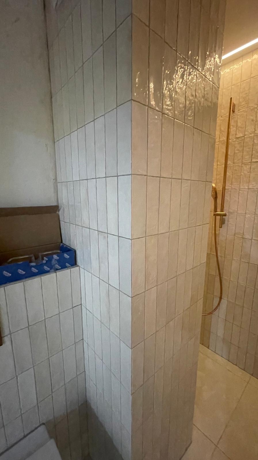 Wykończona ściana w łazience z beżowych, pionowo ułożonych płytek, widoczna kabina prysznicowa z złotą armaturą oraz pudełko z płytkami w rogu ściany.