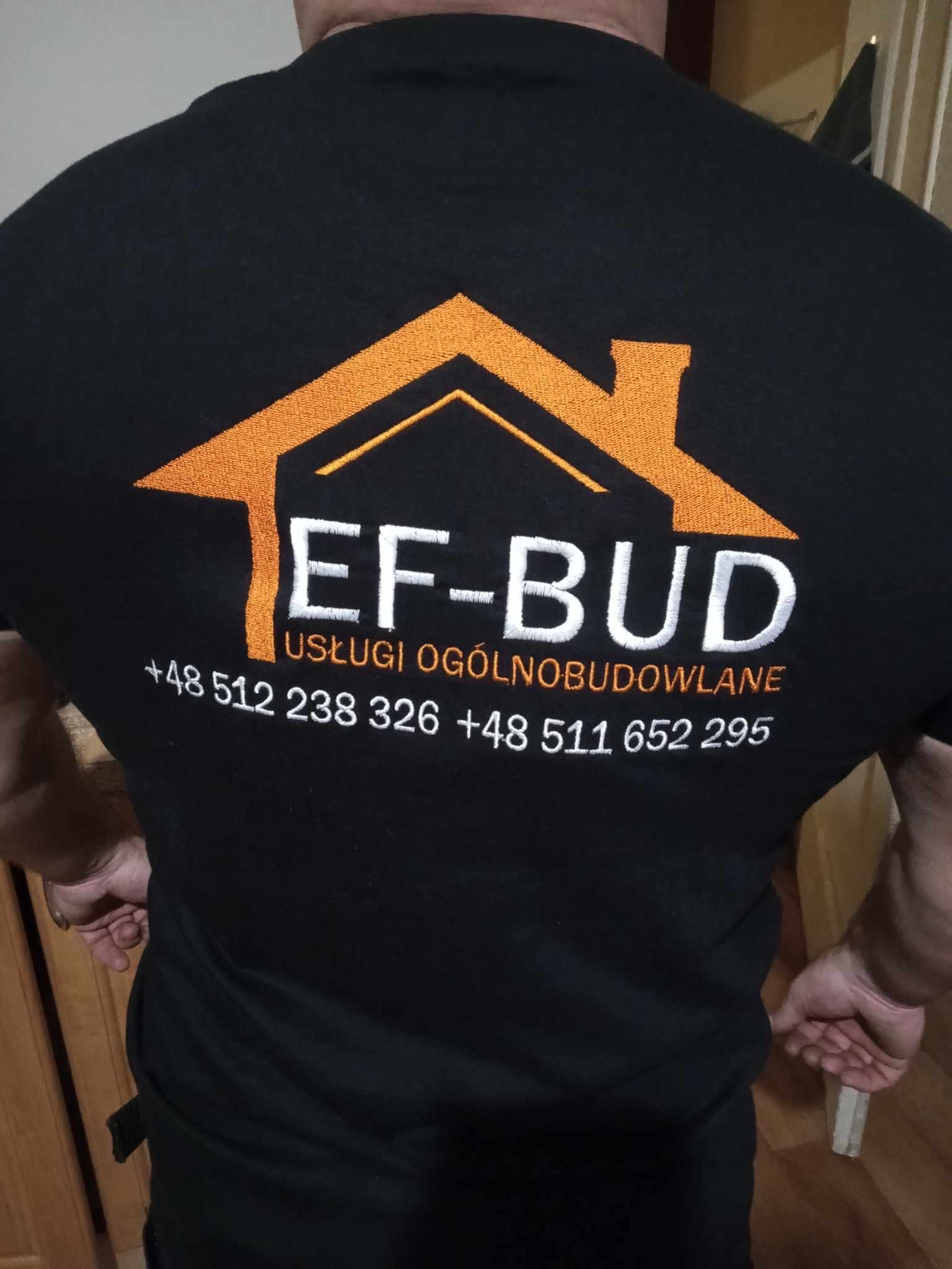 Czarne polo z haftowanym logo firmy EF-BUD, pomarańczowy zarys domu, biały napis EF-BUD, pod spodem USŁUGI OGÓLNOBUDOWLANE i numery telefonów.