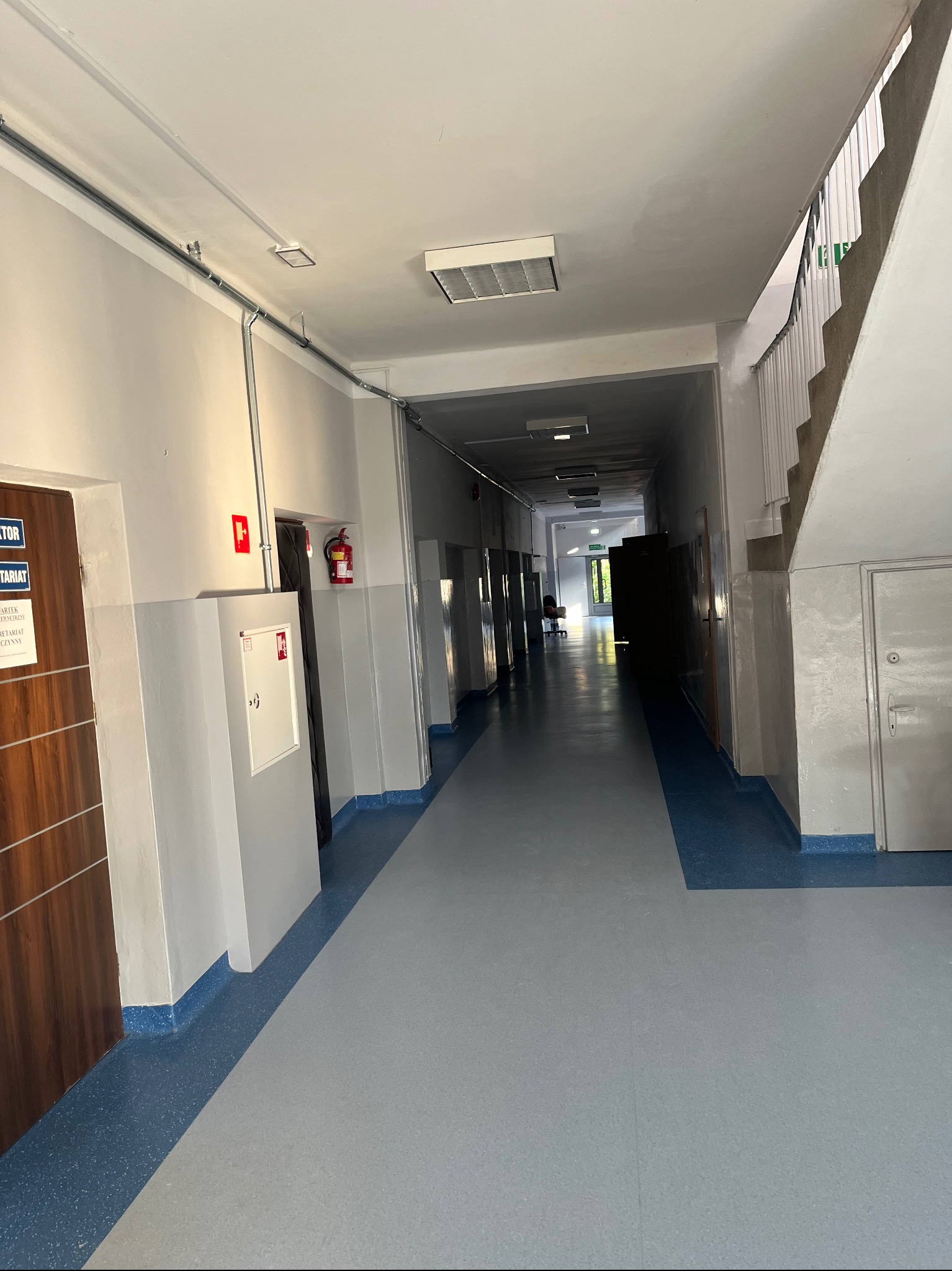 Długi, jasny korytarz w budynku biurowym z szarymi ścianami, niebieską podłogą, widocznymi instalacjami pod sufitem, drzwiami po obu stronach i schodami w oddali.