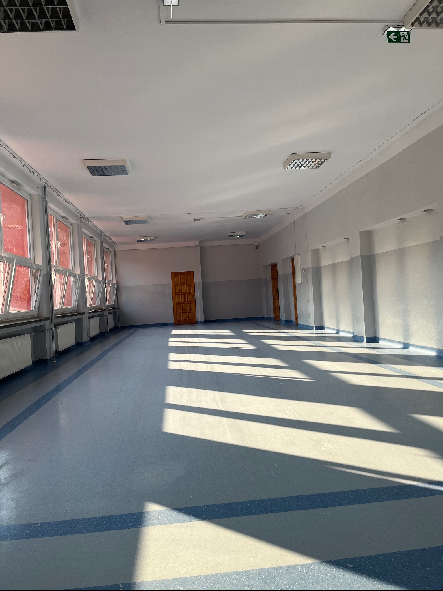 Jasne, przestronne wnętrze korytarza z oknami po lewej stronie, drewnianymi drzwiami na wprost i po prawej stronie, z widocznymi promieniami słonecznymi padającymi na podłogę.