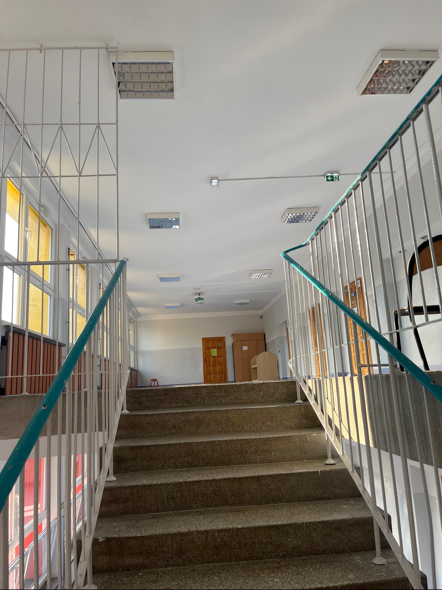 Klatka schodowa w budynku użyteczności publicznej, widok od dołu schodów, z metalowymi barierkami pomalowanymi na biało z turkusowymi akcentami, widoczne okna z żółtymi szybami i sufit z rastrowymi...