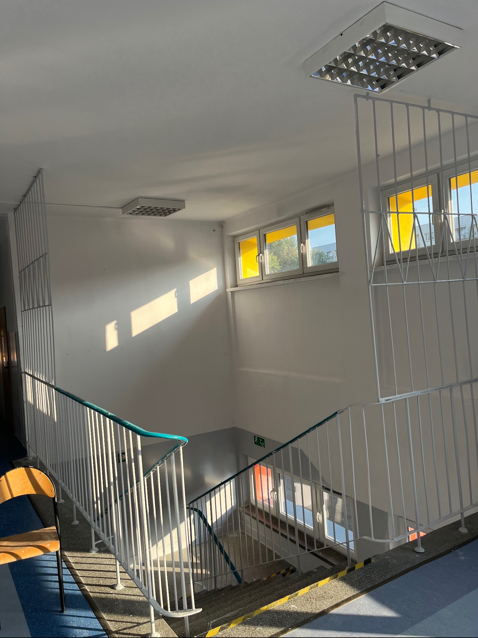 Klatka schodowa w budynku, białe ściany oświetlone promieniami słońca wpadającymi przez okna z żółtymi zasłonami, białe barierki schodów z turkusowymi poręczami, widoczne lampy sufitowe i krzesło.