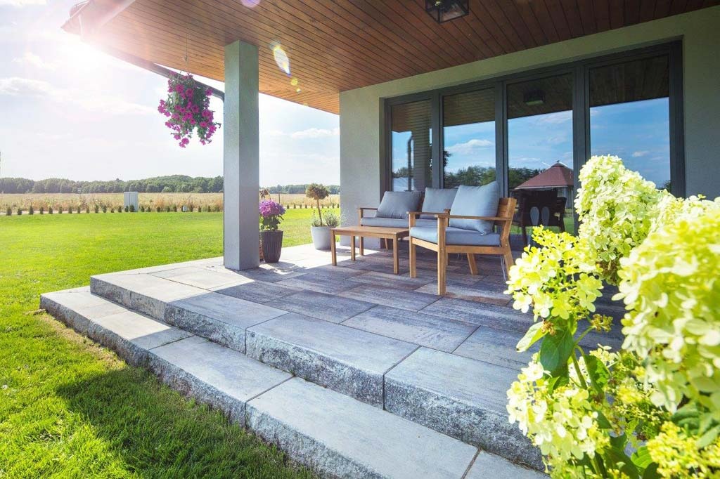 Granitowe schody prowadzą na taras z szarych płyt, na którym stoją drewniane meble ogrodowe, widok na zadbany trawnik i pole w tle.