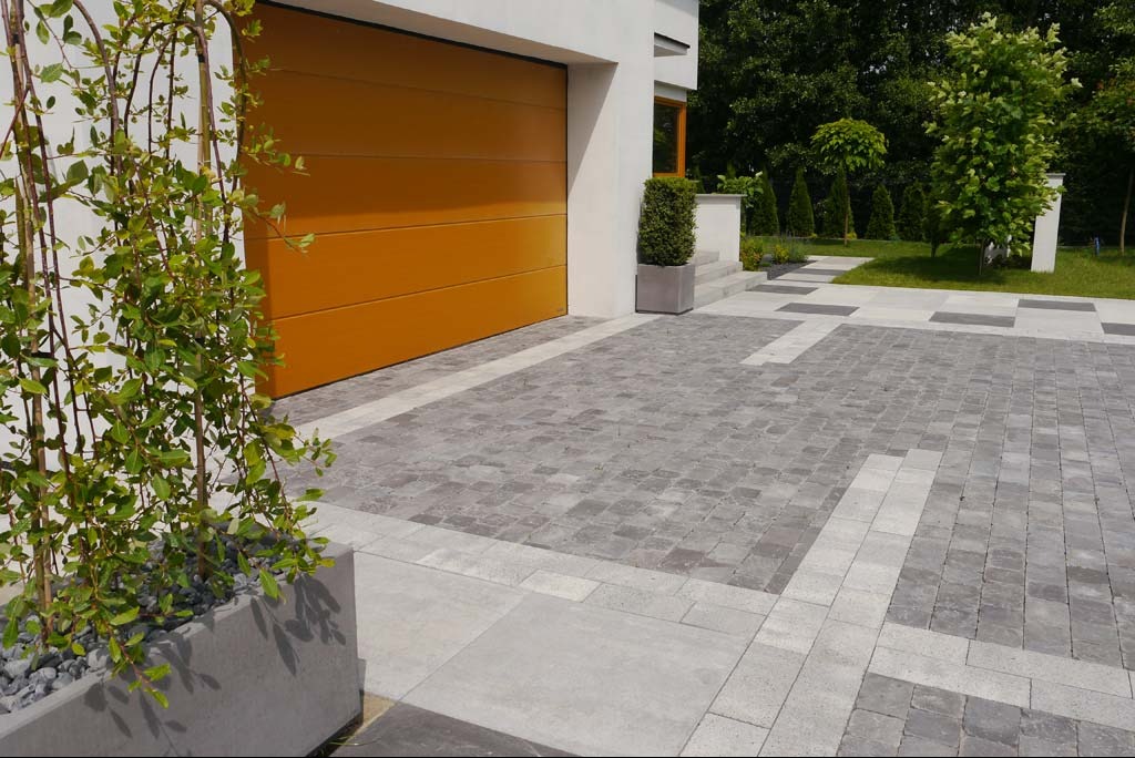 Podjazd z szarej kostki brukowej ułożonej we wzór, obramowany jaśniejszą kostką, z widoczną bramą garażową w kolorze miodowym i roślinnością w donicach.