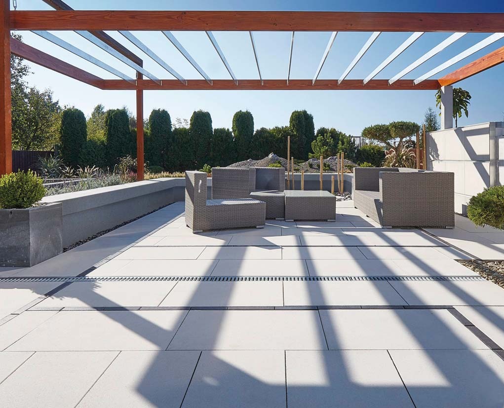 Jasnoszara, prostokątna nawierzchnia tarasu z odpływem liniowym, drewniana pergola z metalowymi poprzeczkami, rattanowe meble ogrodowe i zielony żywopłot w tle.