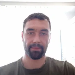 Portret mężczyzny z brodą, ubrany w zieloną koszulkę, z delikatnym łańcuszkiem na szyi, na tle okna z żaluzjami.