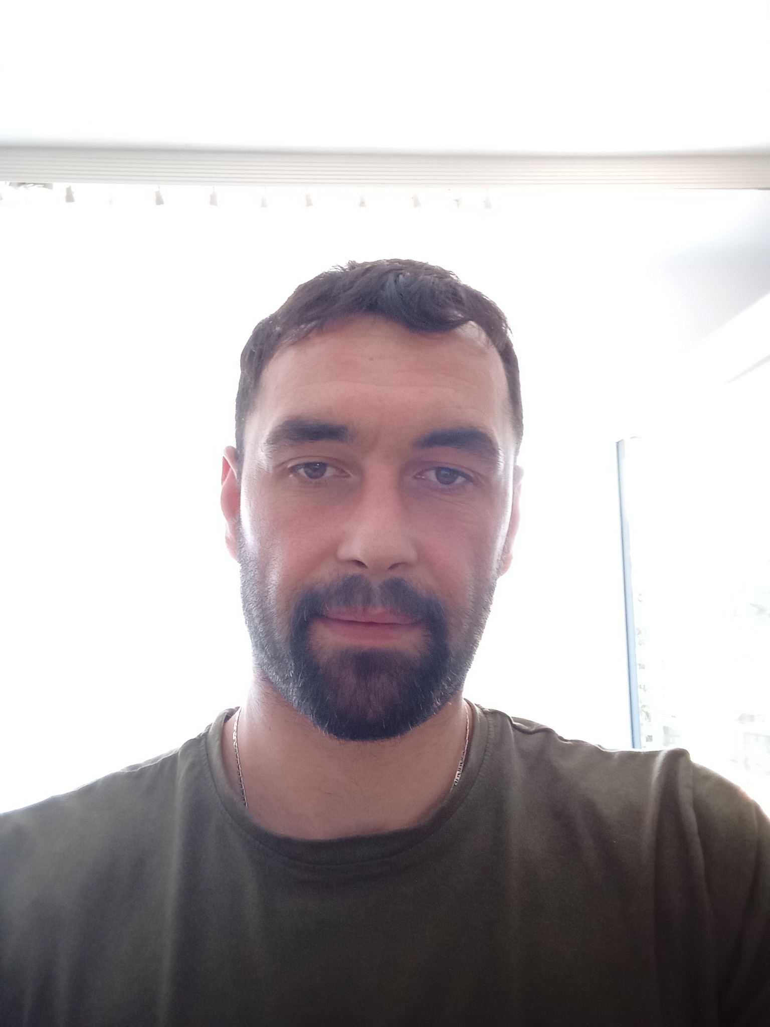 Portret mężczyzny z brodą, ubrany w zieloną koszulkę, z delikatnym łańcuszkiem na szyi, na tle okna z żaluzjami.