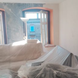 Adam Augustyn - Pokój w trakcie przygotowań do prac wykończeniowych: meble i podłogi zabezpieczone folią, ściany częściowo pokryte szarą warstwą, widoczne okna z pomarańczowymi ramami i budynek za nimi.