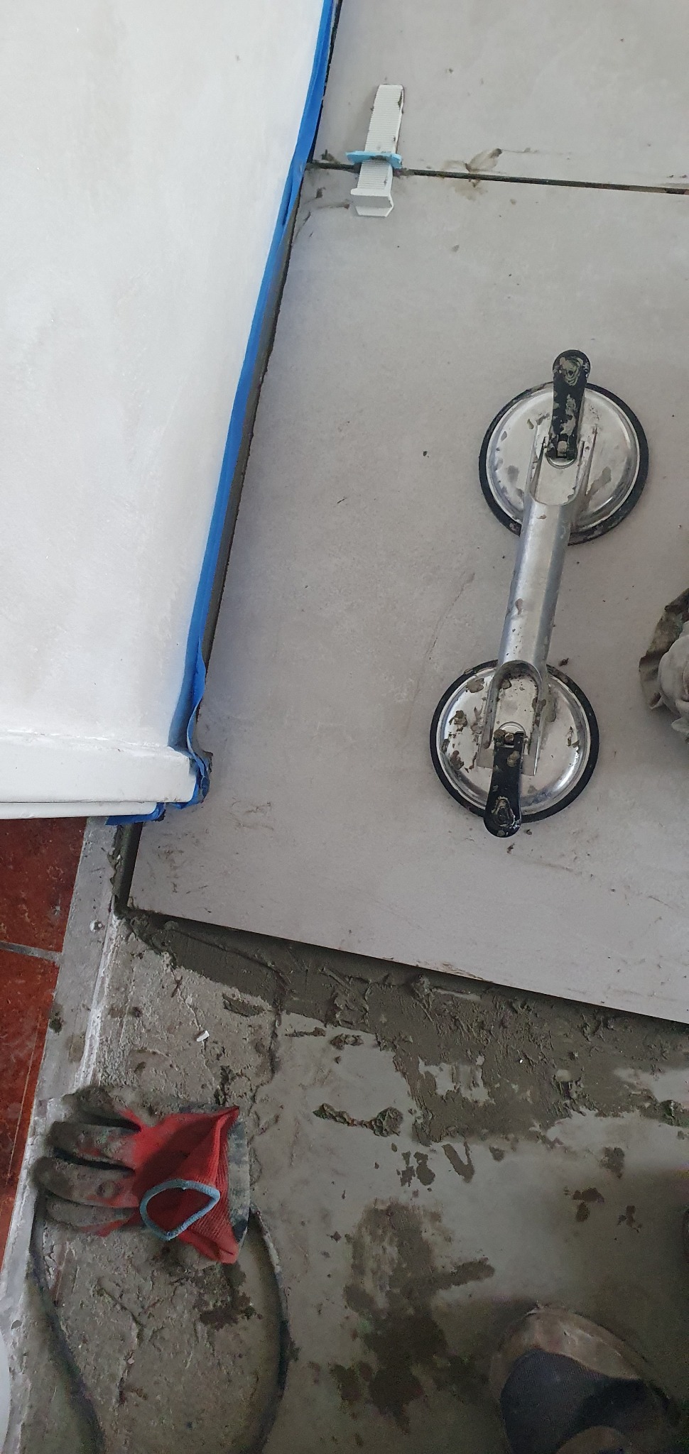 Układanie jasnoszarych płytek podłogowych z użyciem przyssawki do płytek, widoczne krzyżyki dystansowe, niebieska taśma malarska zabezpiecza krawędź wanny, na podłodze resztki zaprawy i robocza...