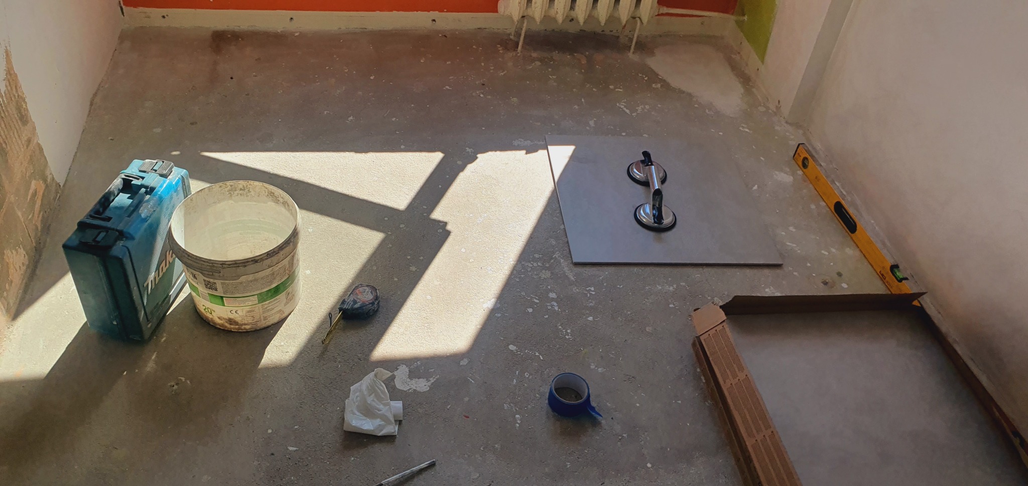 Przygotowanie do układania płytek: płytka z przyssawkami, poziomica, wiadro, skrzynka narzędziowa i zwinięta taśma malarska na surowym podłożu w trakcie remontu.