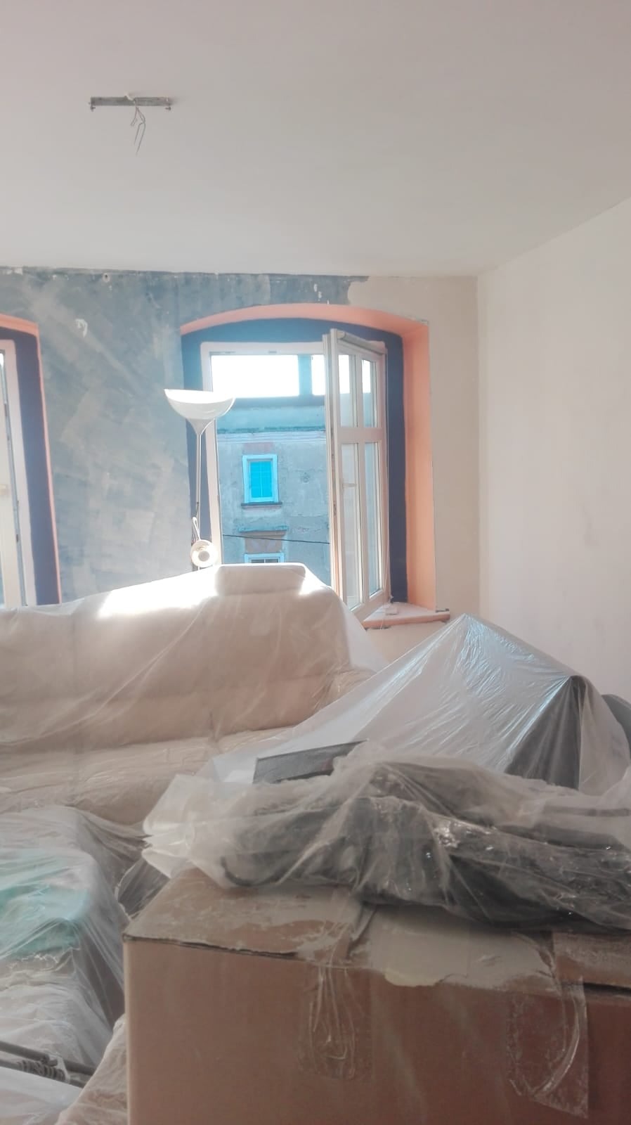 Pokój w trakcie przygotowań do prac wykończeniowych: meble i podłogi zabezpieczone folią, ściany częściowo pokryte szarą warstwą, widoczne okna z pomarańczowymi ramami i budynek za nimi.