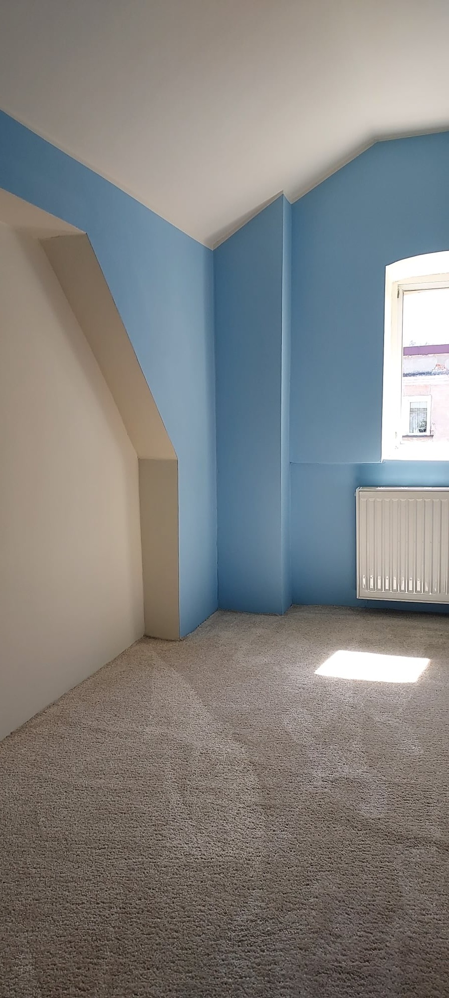 Pomalowane na błękitno wnętrze pokoju z beżową wykładziną, białą ścianą z elementami konstrukcyjnymi i białym grzejnikiem pod oknem.