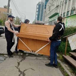 Yaroslav Chetveriakov - Trzech mężczyzn przenosi pianino po chodniku miejskim, zabezpieczone białymi pasami, na tle budynków biurowych i drzew.
