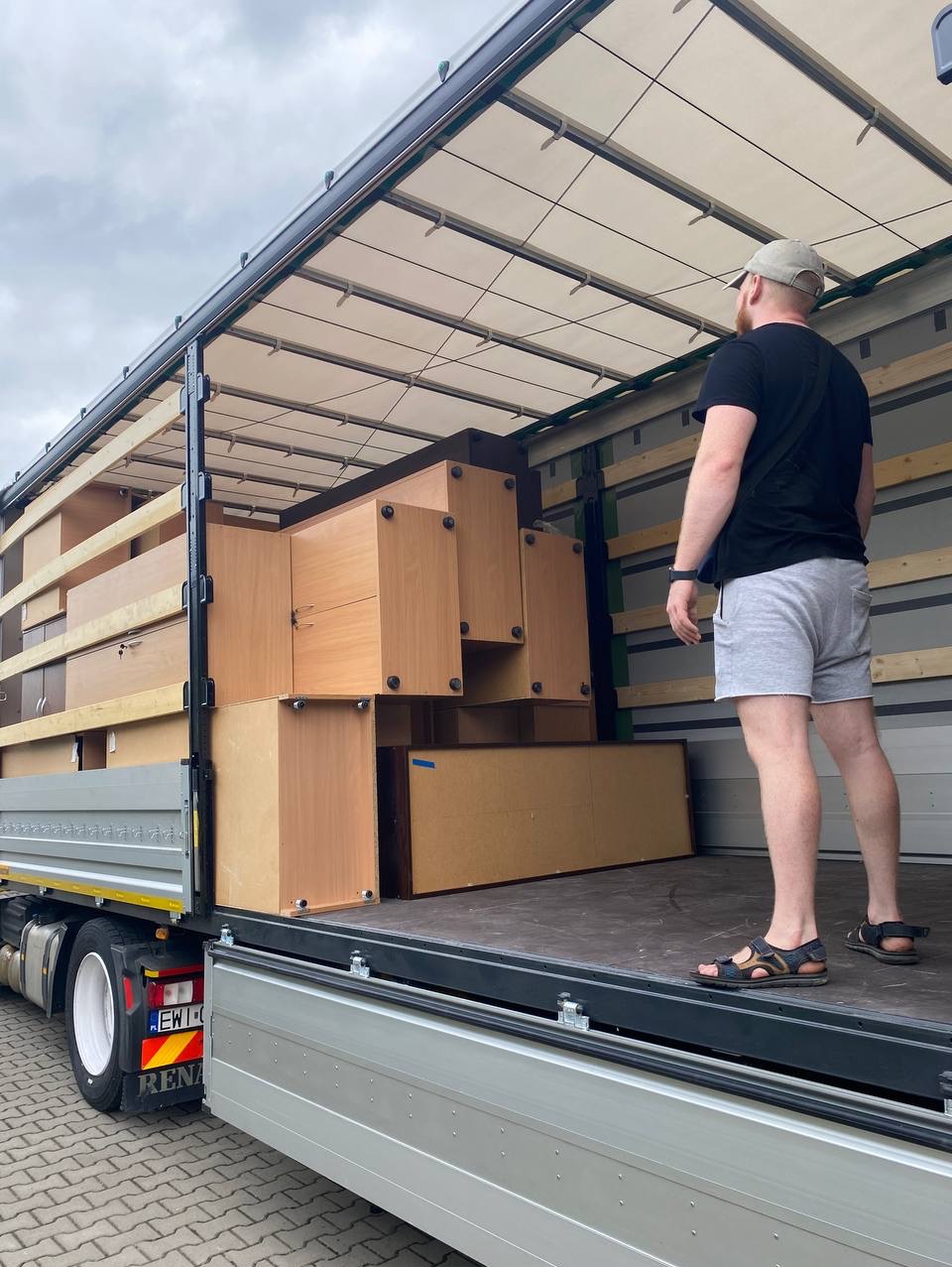 Załadunek mebli skrzyniowych do otwartej naczepy ciężarówki z widocznym mężczyzną w szortach i sandałach stojącym na platformie załadunkowej.