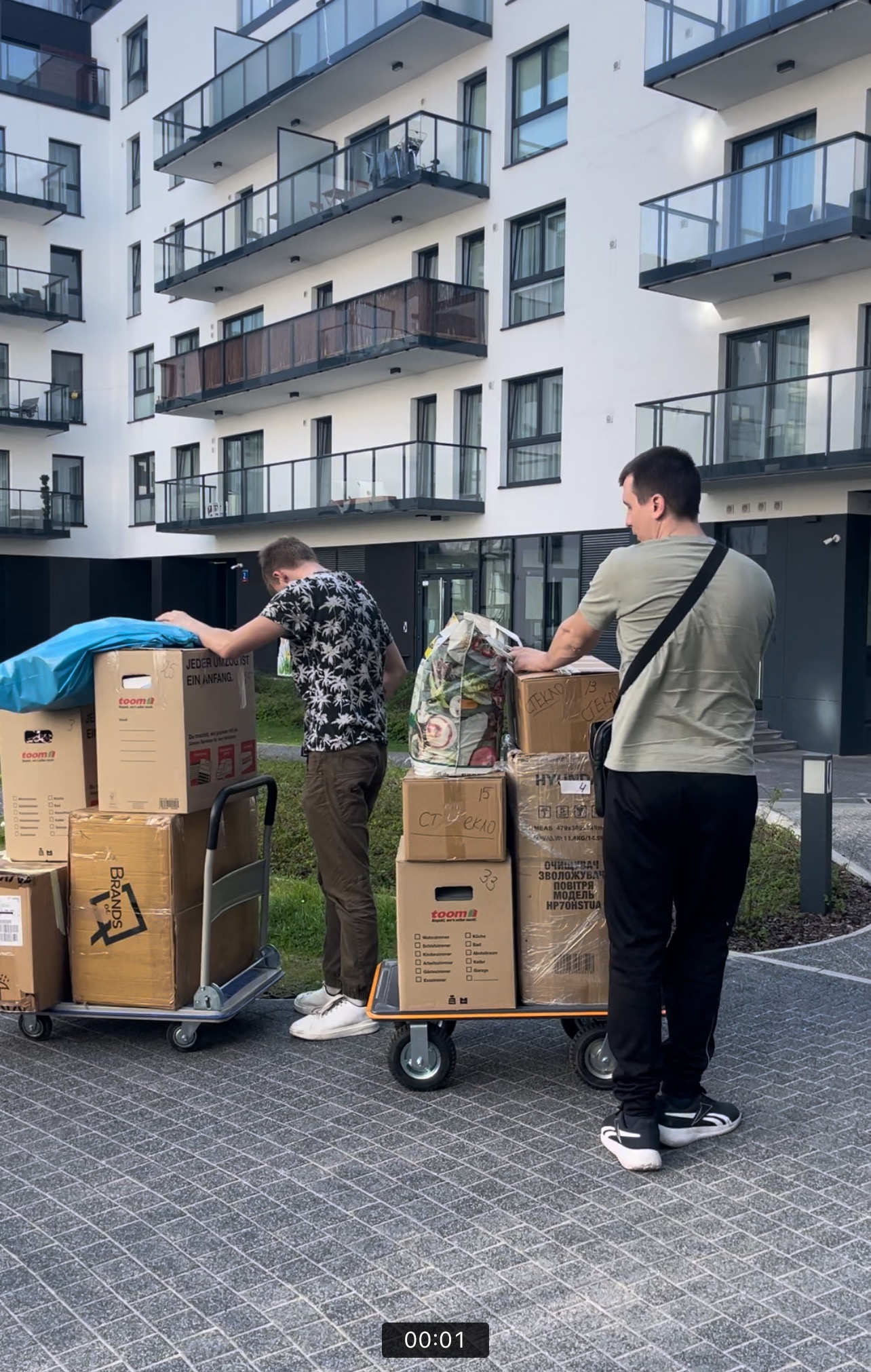 Dwóch mężczyzn transportuje kartony na wózkach przed nowoczesnym budynkiem mieszkalnym; jeden z kartonów oznaczony jako szkło.