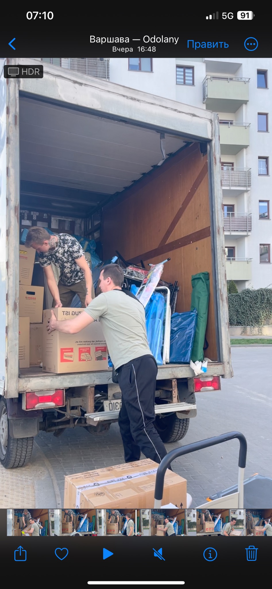 Dwóch mężczyzn ładuje kartony do wnętrza samochodu dostawczego podczas przeprowadzki na tle bloku mieszkalnego w Warszawie, widoczna tablica rejestracyjna samochodu.